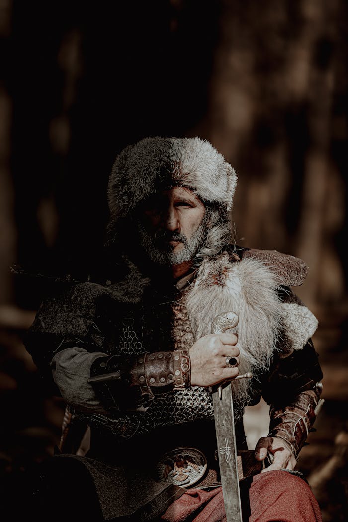 A bearded medieval warrior in fur and armor holding a sword in a dark forest setting.
